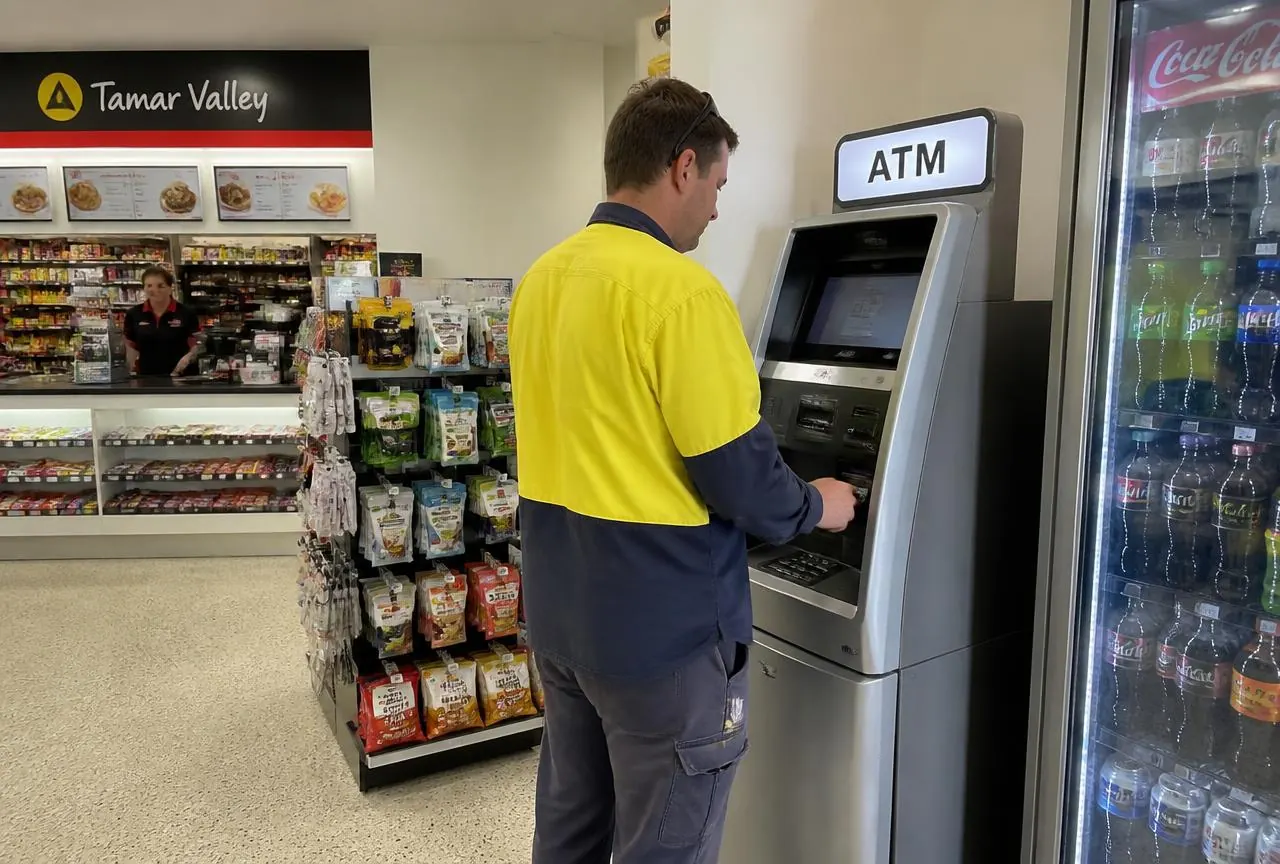 Free-standing ATM machine installed beside the drinks fridges inside an Australian service station shop while a customer withdraws cash